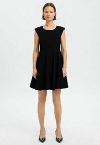 Musta ilman hihoja oleva mekko, jossa on istuva vartalo-osa ja liehuva hame. Valmistettu teksturoidusta kankaasta, jossa on hienovaraisia laskoksia ja pyöreä kaula-aukko.