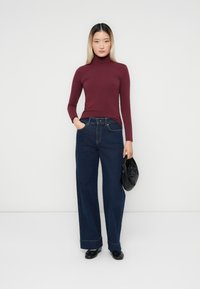 Burgundy pitkähihaisen turtleneck-paita, korkeavyötäröiset tummat farkut leveällä lahkeella, musta laukku kädessä, sileä tekstuuri, minimalistinen tyyli, neutraali tausta.