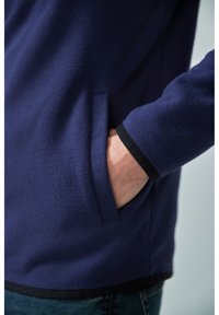Marineblaue Fleecejacke mit seitlicher Tasche, schwarzen Einsätzen an den Bündchen und dem Saum, weicher Textur und einem leicht lockeren Schnitt.