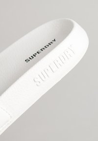 Hvide gummiflip-flops med teksturerede stropper. Har et præget "SUPERDRY" logo på stroppen og en glat sål, minimalistisk design.