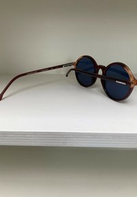 Lunettes de soleil rondes avec des montures en écaille de tortue, verres bleu foncé et branches brunes arborant un logo simple. Légères avec une finition brillante.