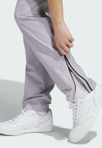 Pantalones deportivos de color púrpura claro con cremalleras laterales negras, combinados con zapatillas blancas que tienen perforaciones y una suela acolchada.