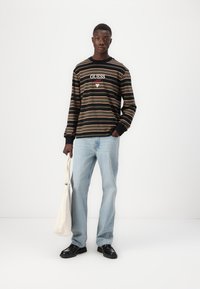 Camisa de manga larga negra y a rayas de cuello redondo con el logo "GUESS", combinada con jeans de color azul claro y zapatos negros, sosteniendo una bolsa de mano beige.