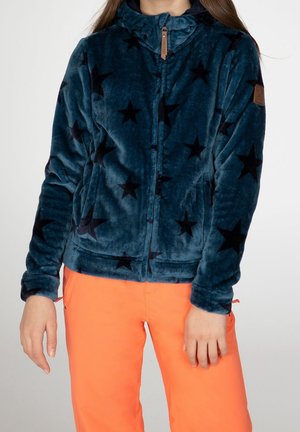 Osoba ubrana w niebieską polarową kurtkę z czarnymi gwiazdkami i jaskrawo pomarańczowe spodnie, widoczna od ramion do bioder.