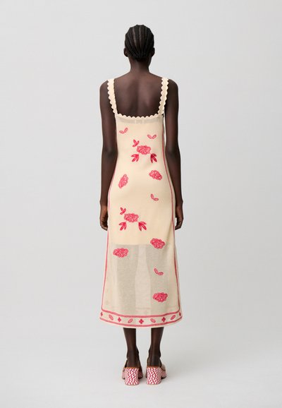 Donna con capelli intrecciati che indossa un vestito midi color crema con ricamo floreale rosso e spalline smerlate, abbinato a tacchi rossi e bianchi con motivo.