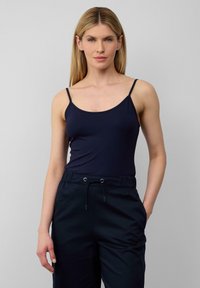 Blonde Frau in einem marineblauen Tanktop und Jogginghose, die mit einer Hand in der Tasche vor einem schlichten hellen Hintergrund steht.