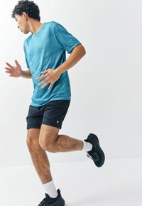 Blå texturerad träningsskjorta och svarta shorts. Svarta löparskor med vita strumpor. Personen ser ut att vara i rörelse, vänd åt sidan.