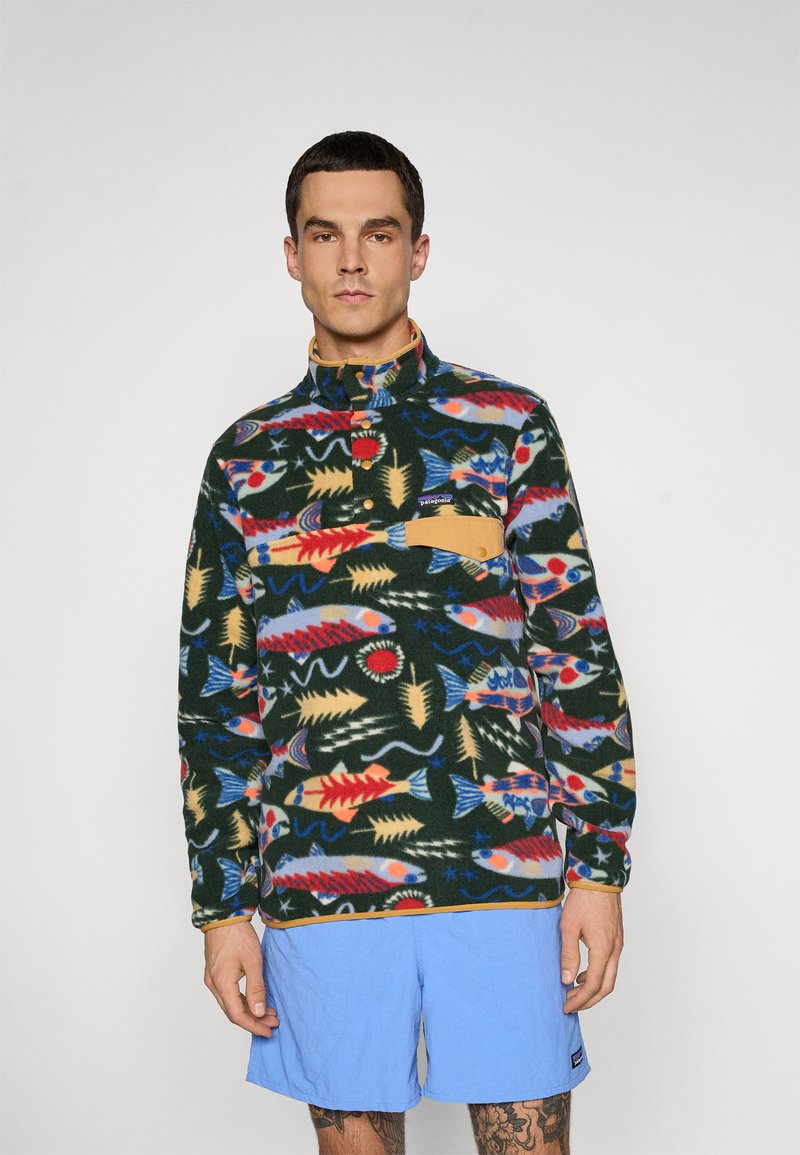 Man in een kleurrijke geblokte fleece pullover met oranje knopen en een zak, gecombineerd met lichtblauwe shorts, staande voor een effen achtergrond.