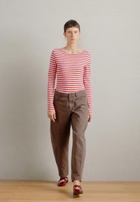 Červené a biele pruhované tričko s dlhým rukávom skombinované s voľnými hnedými nohavicami a červenými sandálmi. Jednoduchý dizajn s uvoľneným strihom a minimálnymi detailmi.