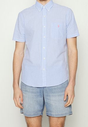 Homme portant une chemise à manches courtes rayée bleu et blanc à boutons et un short en jean bleu clair sur un fond uni.
