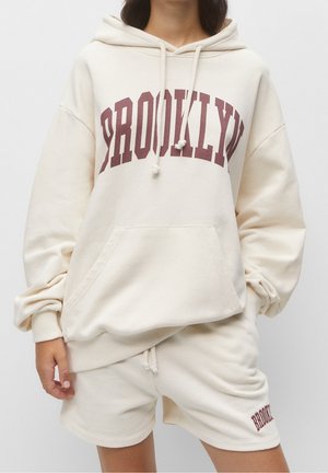 Osoba nosząca kremową bluzę z kapturem z napisem „BROOKLYN” w bordowym kolorze oraz dopasowane szorty z napisem „BROOKLYN” na lewej nodze.