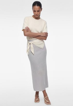 Frau mit dunklen, zurückgebundenen Haaren, die ein cremefarbenes, kurzärmeliges Oberteil mit Seitenschleife, einen hellgrauen langen Rock und beige Sandalen mit Riemchen trägt.