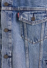 Giubbotto in denim azzurro chiaro con bottoni a pressione in metallo, tasca sul petto e cuciture arancioni a contrasto. Tessuto testurizzato con una finitura sbiadita.