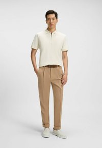 Gräddfärgad polo-shirt med dragkedja i kragen, i kombination med beiga veckade byxor och vita slip-on-skor med ett strukturerat mönster.