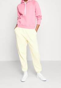 Rosa huvtröja med dragsko och framficka, tillsammans med gula sweatpants som har elastisk midja och muddar, bärs med vita sneakers.