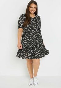 Musta kukkamekko lyhyillä hihoilla ja kerroksellisella hameella. Ominaisuuksia ovat pyöreä kaula-aukko, hajallaan olevat valkoiset ja keltaiset kukkatyylit, yhdistettynä valkoisiin tennareihin.