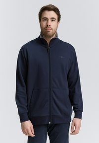 Giacca zip-up blu navy realizzata in tessuto morbido, con colletto alto, due tasche frontali e polsini elastici, dotata di un discreto dettagli logo sul petto.