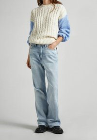 Kerman värinen palmikkoneulepaita, jossa on siniset resorit hihansuissa, yhdistetty vaaleansinisillä kulutetuilla suoralahkeisilla farkuilla ja mustilla massiivisilla kengillä.