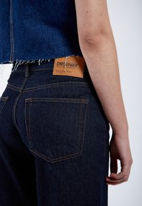 Jeans de algodón denim azul oscuro con costuras naranjas. Presenta una etiqueta de cuero con "DRDENIM" en la cinturilla trasera y bolsillos traseros estándar.