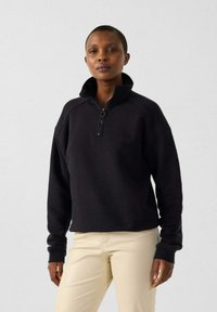 Sweat-shirt noir à col zippé mi-hauteur, avec des poignets côtelés et une coupe décontractée, associé à un pantalon beige. Texture douce, design minimal.
