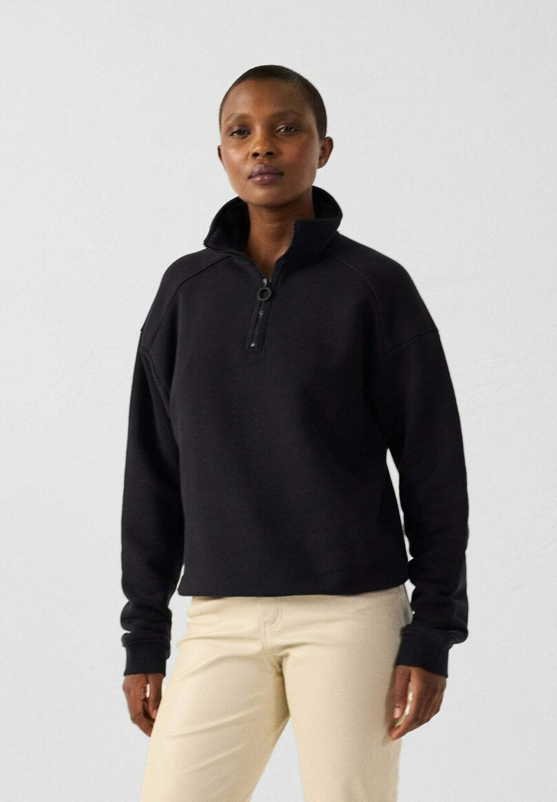 Sweat-shirt noir à col zippé mi-hauteur, avec des poignets côtelés et une coupe décontractée, associé à un pantalon beige. Texture douce, design minimal.