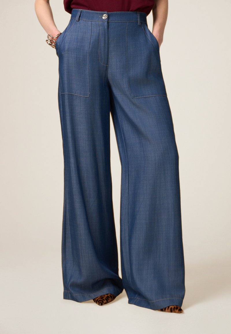 Pantaloni a gamba larga in tessuto blu scuro con una texture liscia, dotati di tasche frontali e chiusura con bottone. Accentuati da una scarpa con stampa leopardo.