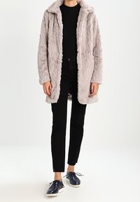 Ljust grå, texturerad fuskpälsjacka med krage, bärs över en svart skjorta, kombinerad med svarta byxor och mörkblå skor.