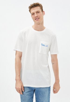 Witte katoenen t-shirt met korte mouwen, ronde halslijn en een borstzak met blauwe tekst. Gecombineerd met lichtblauwe denim jeans.