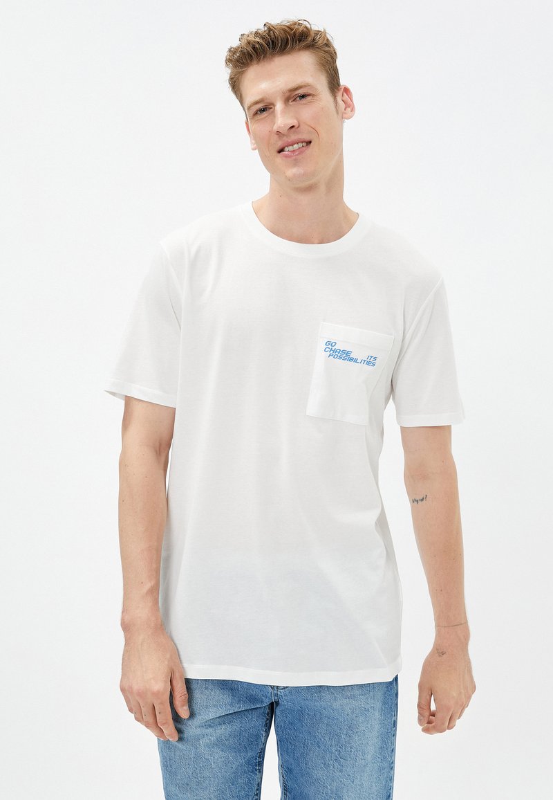 Camiseta de algodón blanca de manga corta, con escote redondo y un bolsillo en el pecho que presenta texto azul. Combinada con jeans de mezclilla azul claro.