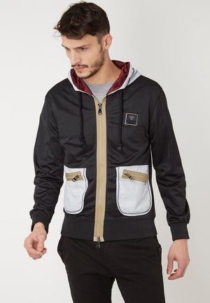 Chaqueta negra con cremallera y capucha gris, con forro burdeos, que cuenta con dos bolsillos blancos con cremalleras y detalles de cremallera dorados.