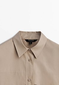 Chemise beige à boutons avec col classique, deux poches avant et texture de tissu lisse. Comprend des boutons assortis et une étiquette dans l'encolure.