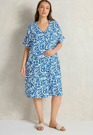 Donna in piedi che indossa un vestito blu e bianco con motivo a foglie, lungo fino al ginocchio, con maniche corte e sandali beige a ciabatta.