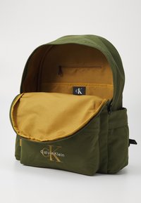 Vihreä kangasreppu kultaisella sisällä, vetoketjullinen tasku ja Calvin Klein -logo. Ominaisuuksiin kuuluu pyöristetty yläosa ja kestävä ommel.
