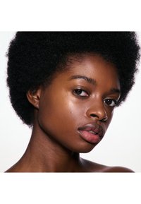 Gros plan d'une personne avec des cheveux afro naturels, une peau lisse, un anneau nasal et une expression faciale neutre sur un fond clair.