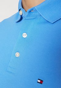 Světle modrá polo tričko vyrobené z texturované tkaniny, s malým logem a dvěma bílými knoflíky na límci.