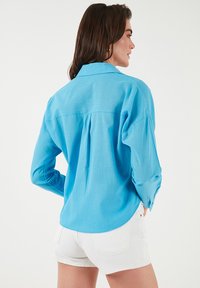 Light blue long-sleeve shirt with a button-down collar and pleated back detail. Paired with white shorts, featuring a curved hemline.