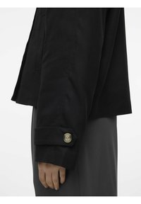 Giacca nera con silhouette accorciata, tessuto testurizzato e polsini abbottonati di un tono di ottone opaco. Indossata con pantaloni grigi.