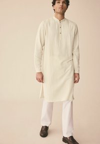 Cremefarbenes Kurta mit strukturierter Oberfläche, das einen Stehkragen und drei Knöpfe hat. Kombiniert mit weißen Hosen und braunen Schuhen.