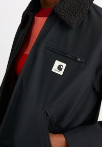 Zwarte jas met een fleece kraag, ritszak en een wit logo-label. Het materiaal lijkt glad en stevig met een getailleerde pasvorm.