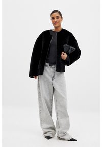 Falošná kožušinová čierna bomber bunda s krátkym golierom, v kombinácii s voľnými svetlošedými džínsami. Modelka drží čierny clutch.