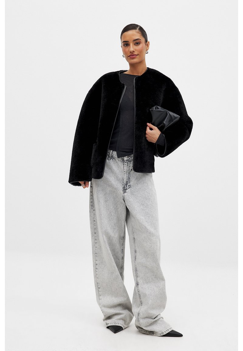 Falošná kožušinová čierna bomber bunda s krátkym golierom, v kombinácii s voľnými svetlošedými džínsami. Modelka drží čierny clutch.