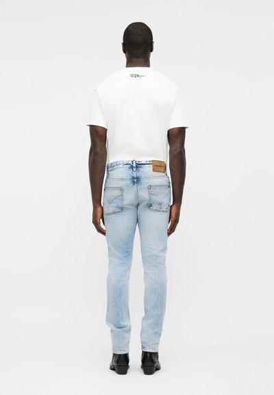 Hombre vistiendo una camiseta blanca de manga corta y jeans azul claro descoloridos, de pie de espaldas sobre un fondo blanco liso.