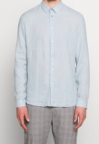 Uomo vestito con una camicia a maniche lunghe azzurra chiara e pantaloni a quadri grigi, in piedi di fronte a uno sfondo semplice.