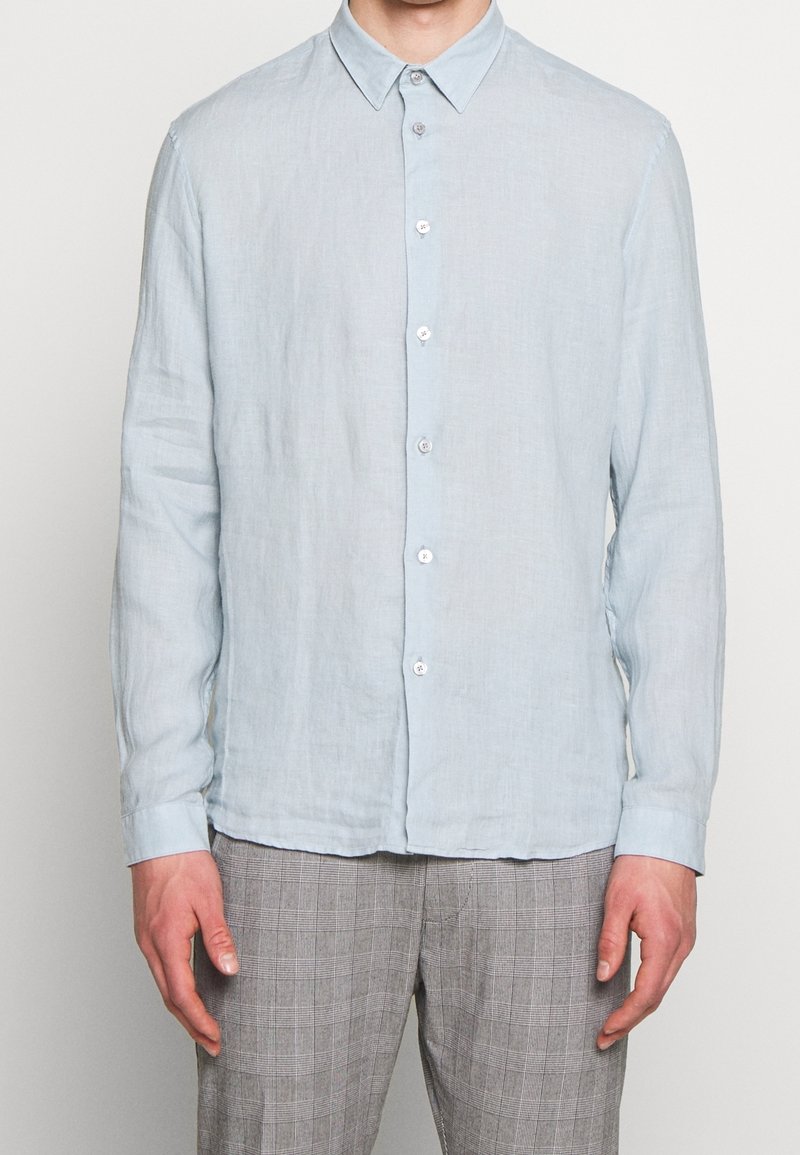 Uomo vestito con una camicia a maniche lunghe azzurra chiara e pantaloni a quadri grigi, in piedi di fronte a uno sfondo semplice.