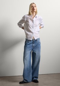 Camicia bianca a maniche lunghe con sottili righe, abbinata a jeans a gamba larga di colore azzurro chiaro. Il modello è in piedi con le mani sui fianchi, indossando calzature nere.