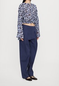 Blouse à motif floral bleu et blanc, nouée à la taille. Pantalon large bleu, coupe ajustée avec plis à l'avant. Chaussures plates noires.