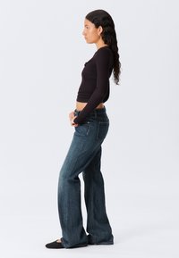 Zwarte lange mouwen top en donkerblauwe flared jeans. De jeans hebben een klassieke snit met subtiele stiksels. Model staat in een zijprofiel.