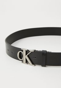 Ceinture en cuir noir avec une boucle en métal argenté « CK », dotée d'une texture lisse et de coutures contrastantes le long des bords.