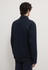 Giacca blu navy con una texture liscia, design con colletto e dettagli in bottone sui polsini; presenta una silhouette pulita e minimalista.