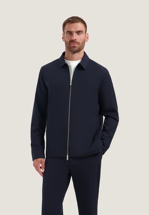 Uomo con giacca blu navy con zip sopra una camicia bianca e pantaloni blu navy abbinati, in piedi davanti a uno sfondo chiaro e uniforme.